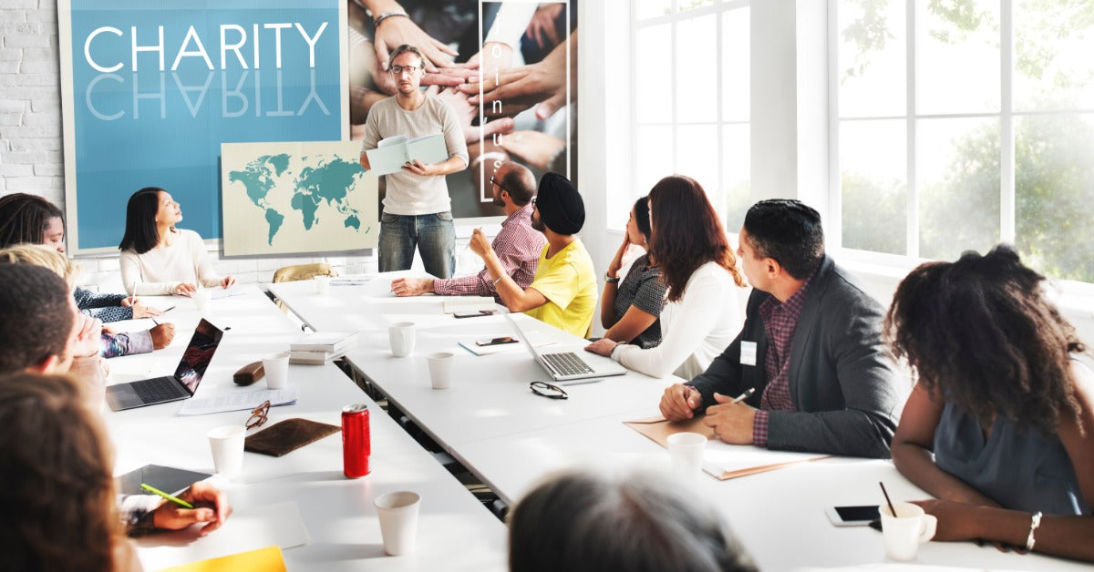 A man standing beside a world map presenting to a group of people seated around a long conference table in a bright room.